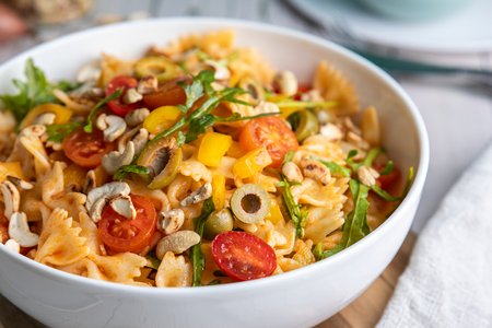 Mediterraner Nudelsalat mit Farfalle, Oliven, Tomaten, Rucola in einer weißen Salatschüssel auf einem Tisch