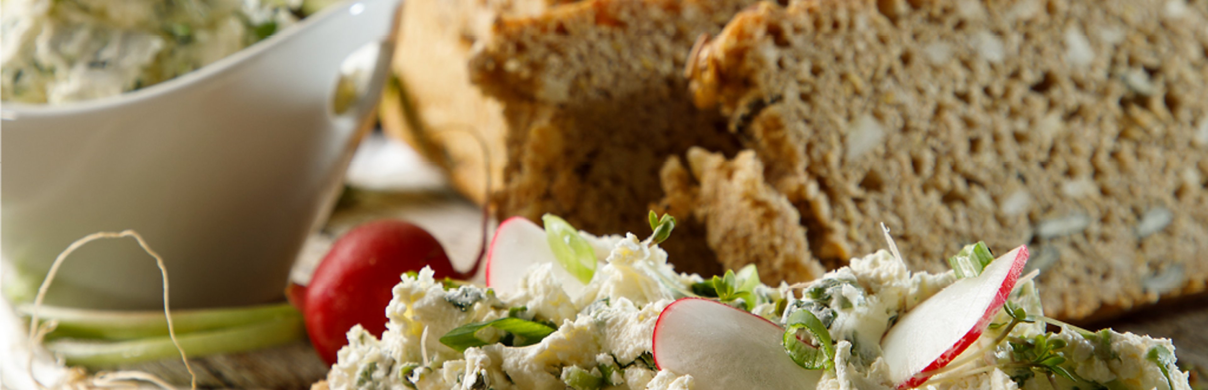 Eine Scheibe Brot auf einem Holzbrett mit selbstgemachten Kräuterfrischkäse und Radieschen belegt. Im Hintergrund ein paar weitere Scheiben Brot und eine weiße Schale mit dem Kräuterdip