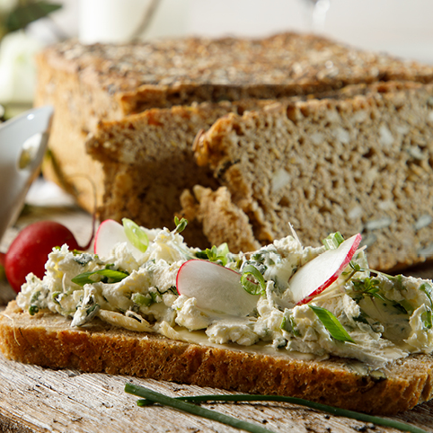 Eine Scheibe Brot auf einem Holzbrett mit selbstgemachten Kräuterfrischkäse und Radieschen belegt. Im Hintergrund ein paar weitere Scheiben Brot und eine weiße Schale mit dem Kräuterdip
