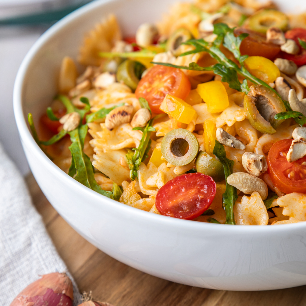 Mediterraner Nudelsalat mit Farfalle, Oliven, Tomaten, Rucola in einer weißen Salatschüssel auf einem Tisch