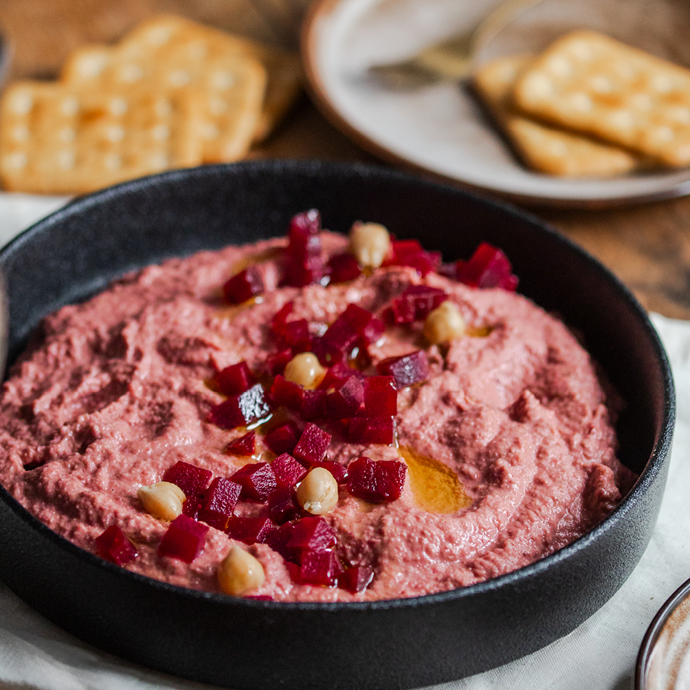 Rote Bete Hummus, angerichtet auf einem tiefen schwarzen Teller, garniert mit kleinen Rote Bete Stückchen.